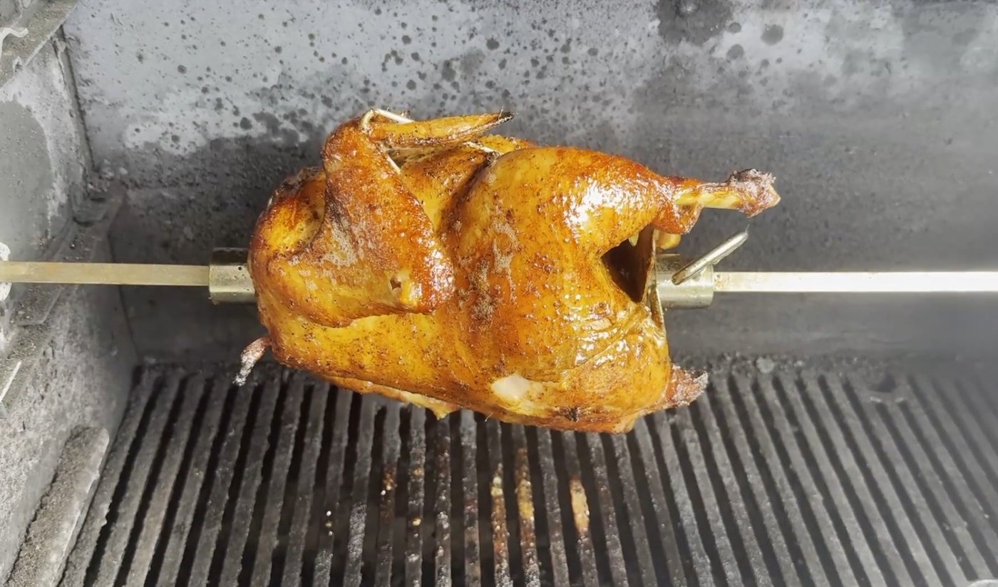 Whole chicken with golden crispy skin turning on a rotisserie over the grill